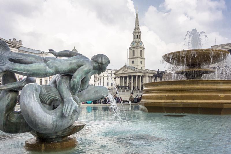 Statue of a mermaid with dolphins in the fountains of Trafalgar Square, with a statue of King George IV on his horse and St Martin in the Fields church in the background, London, UK