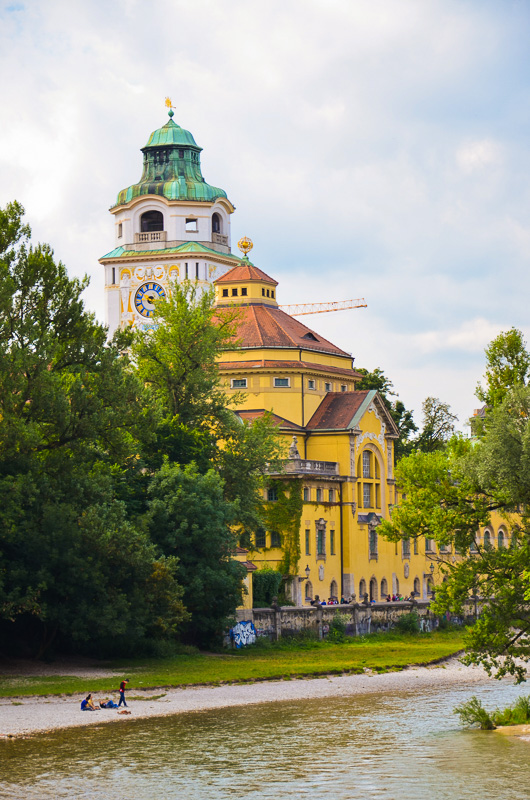 Yellow art mouveau-style building with white tower behhind it beside the river