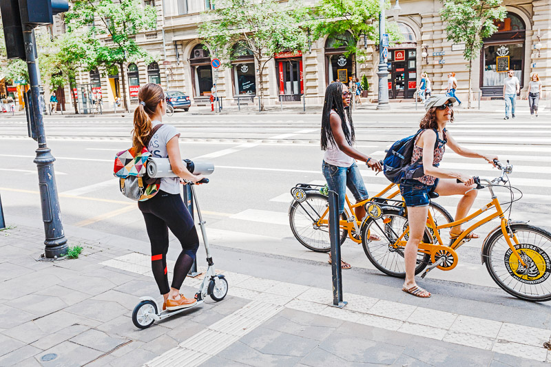  A woman on a kick scooter near the pedestrian crossing is waiting for the bicyclists to pass by