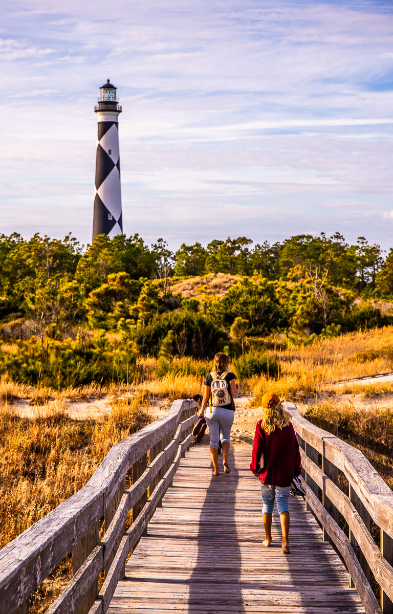 woman and girl walking on a wooden boardwalk with lighthouse in distance