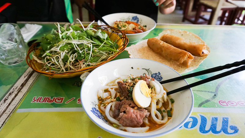 bowl of vietnamese mi quang on table with spring rolls