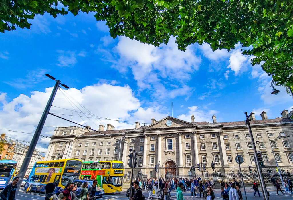 People crossing a street in front of buses and a college in Dublin