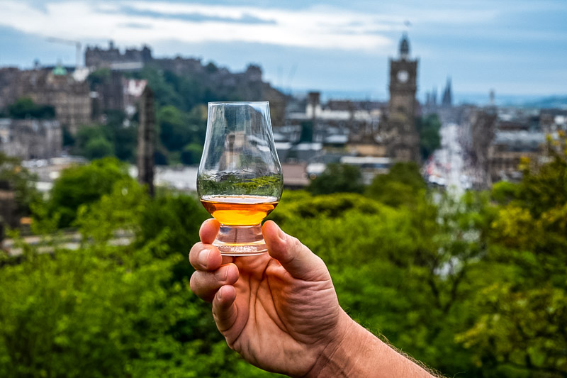 Hand holding glass of single malt scotch whisky and view from Calton hill to park and old parts of Edinburgh city in rainy summer day, Scotland, UK