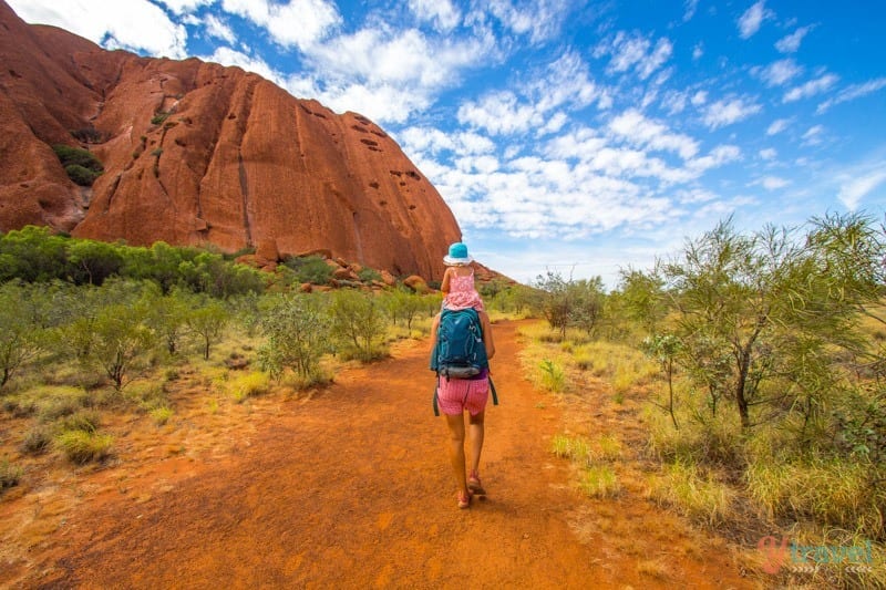 woman carrying child on shoulders walking beside uluru