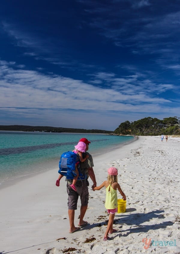 man holding girls hand with toddler in hiking carrier as they walk on the beawch