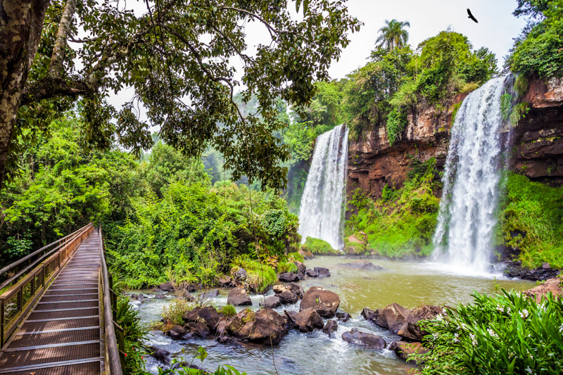 wooden walkways laid in front of two powerful fairy waterfalls from Iguazu Falls fall into a small quiet pond.