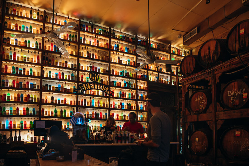 back bar filled with bottles and people standing at bar  beside ouzo barrels