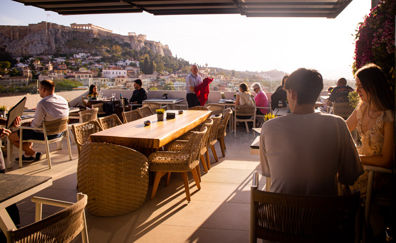 people sitting at tables on rooftop bar with view of acropolis