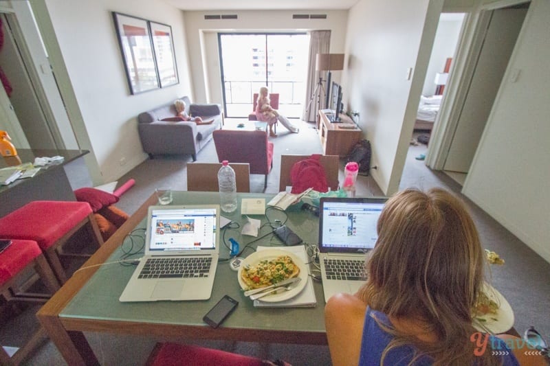 caz working on computer while girls watch tv in hotel apartment