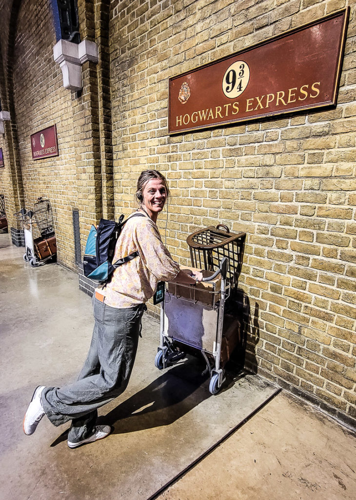 woman posing for a photo in front of a brick wall while holding onto a luggage cart