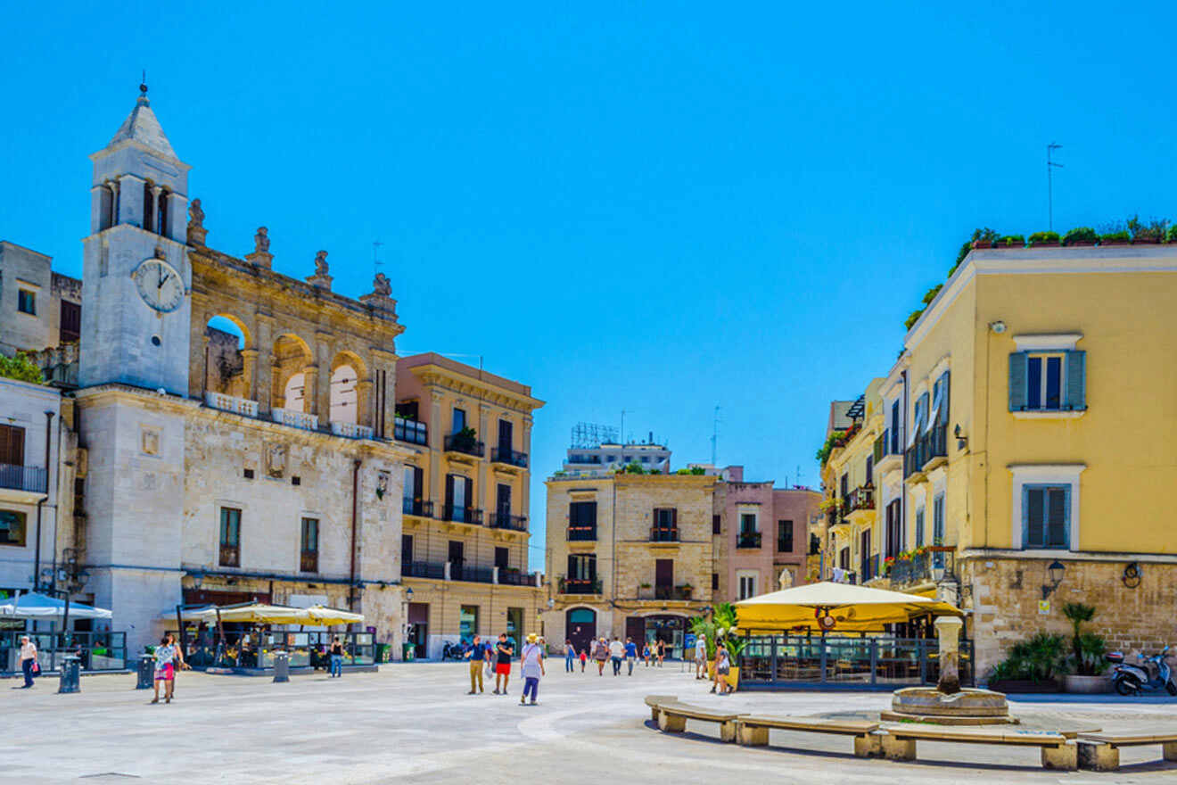 A square in a town with a clock tower and other buildings and people walking around