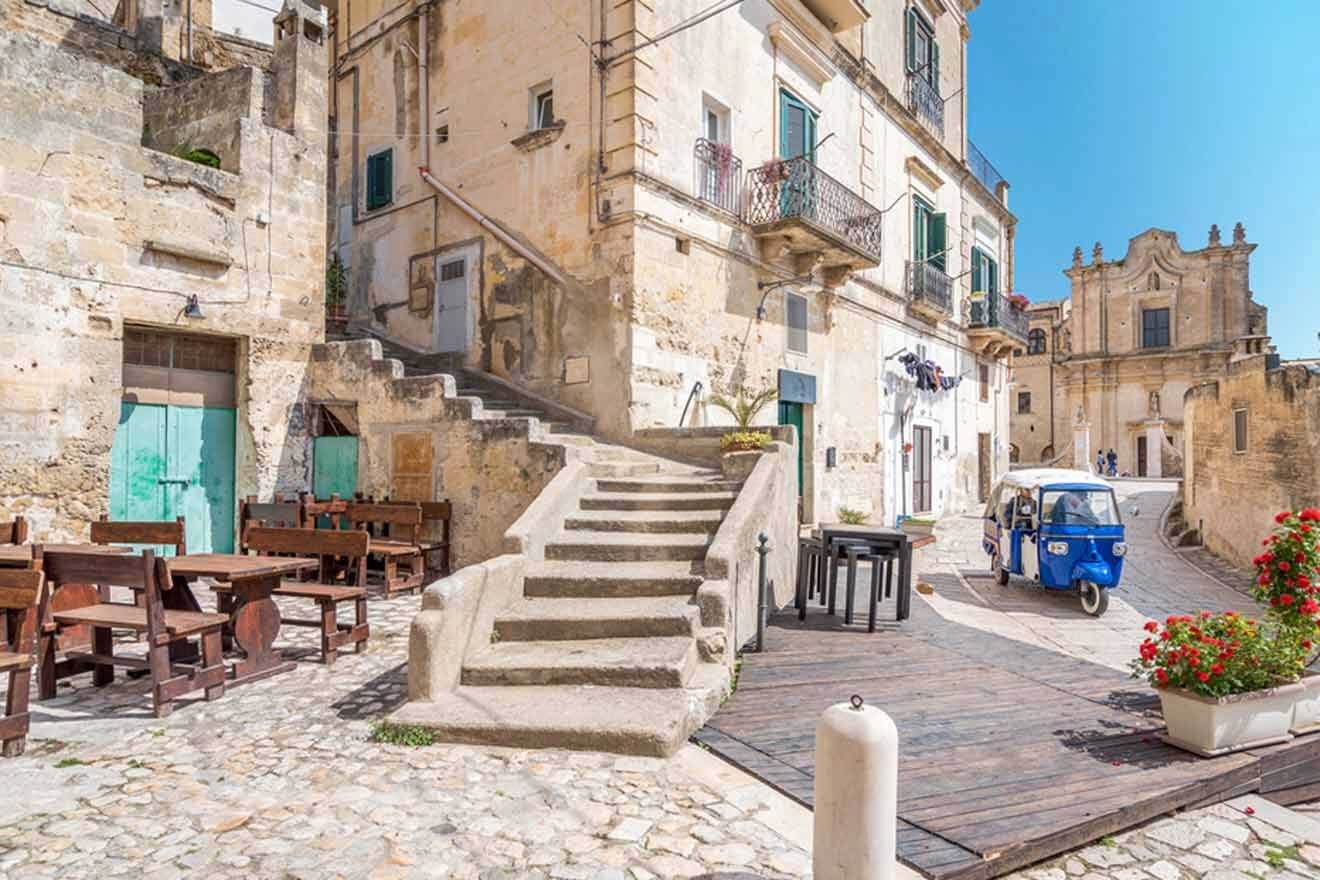 a blue vehicle parked in front of a stone building with stairs leading up to it and wooden benches and tables