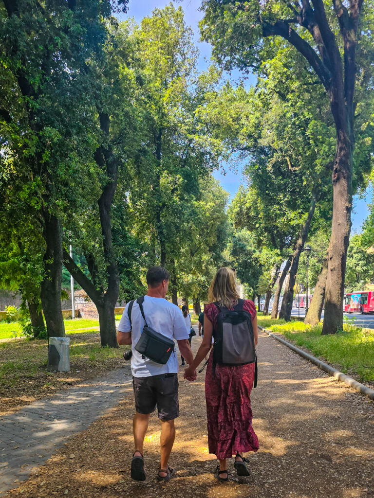 caz and craig holding hands walking through tree lined street in borghese gardens