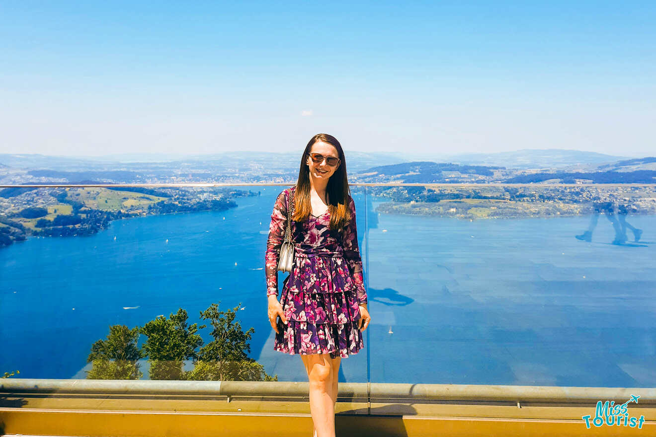 A woman in a floral dress standing on a balcony overlooking a lake.