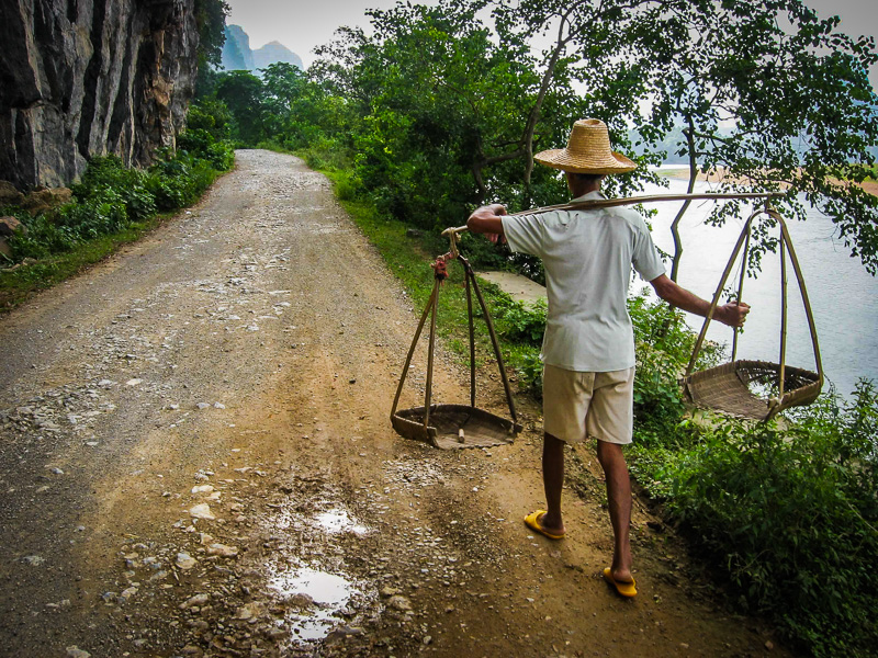 man carrying baskets on his shoulders on country road