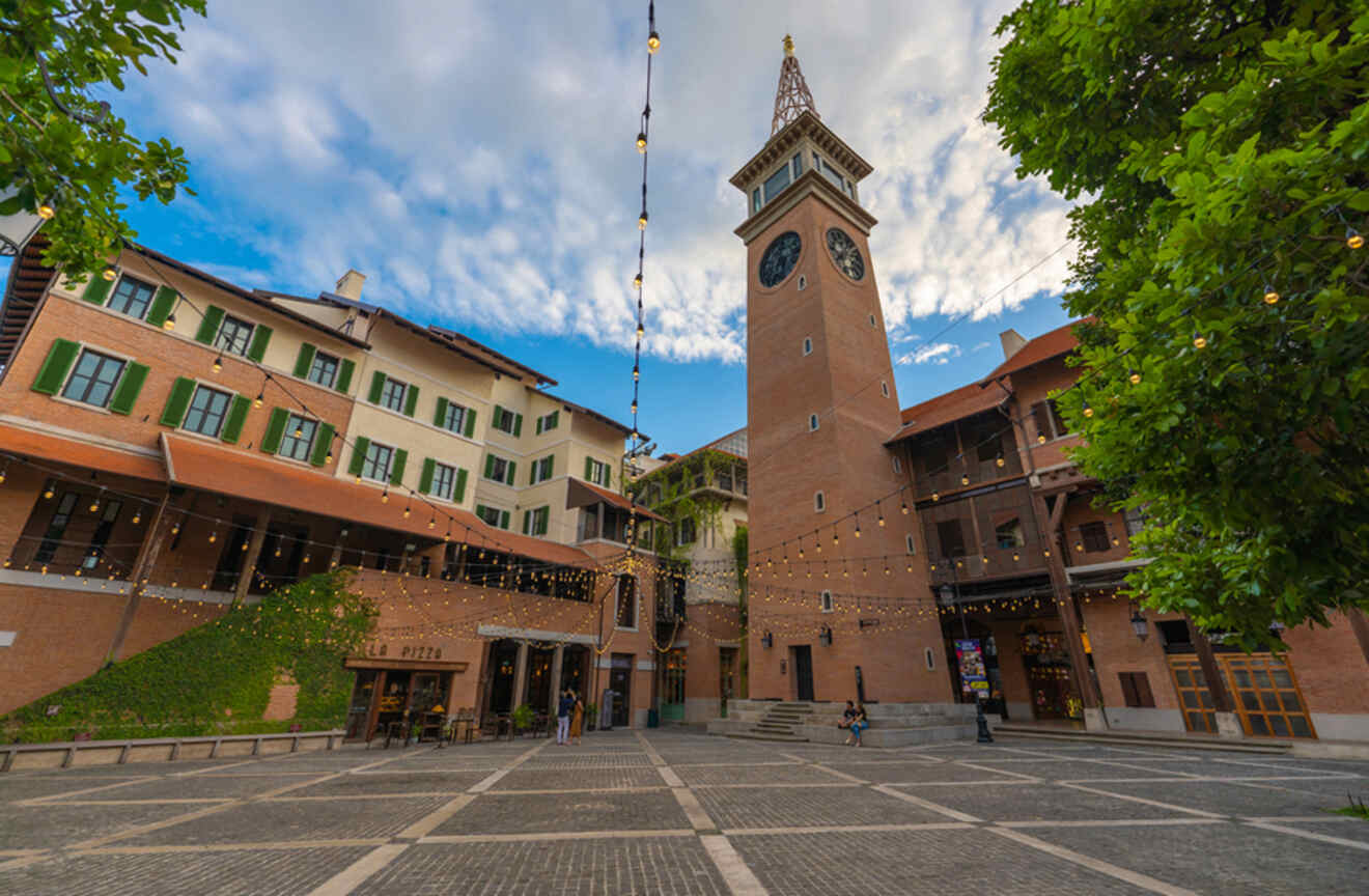 A charming European-style square with a clock tower, warmly lit bistros, and festoon lighting, giving a cozy ambiance in a modern Chiang Mai community.