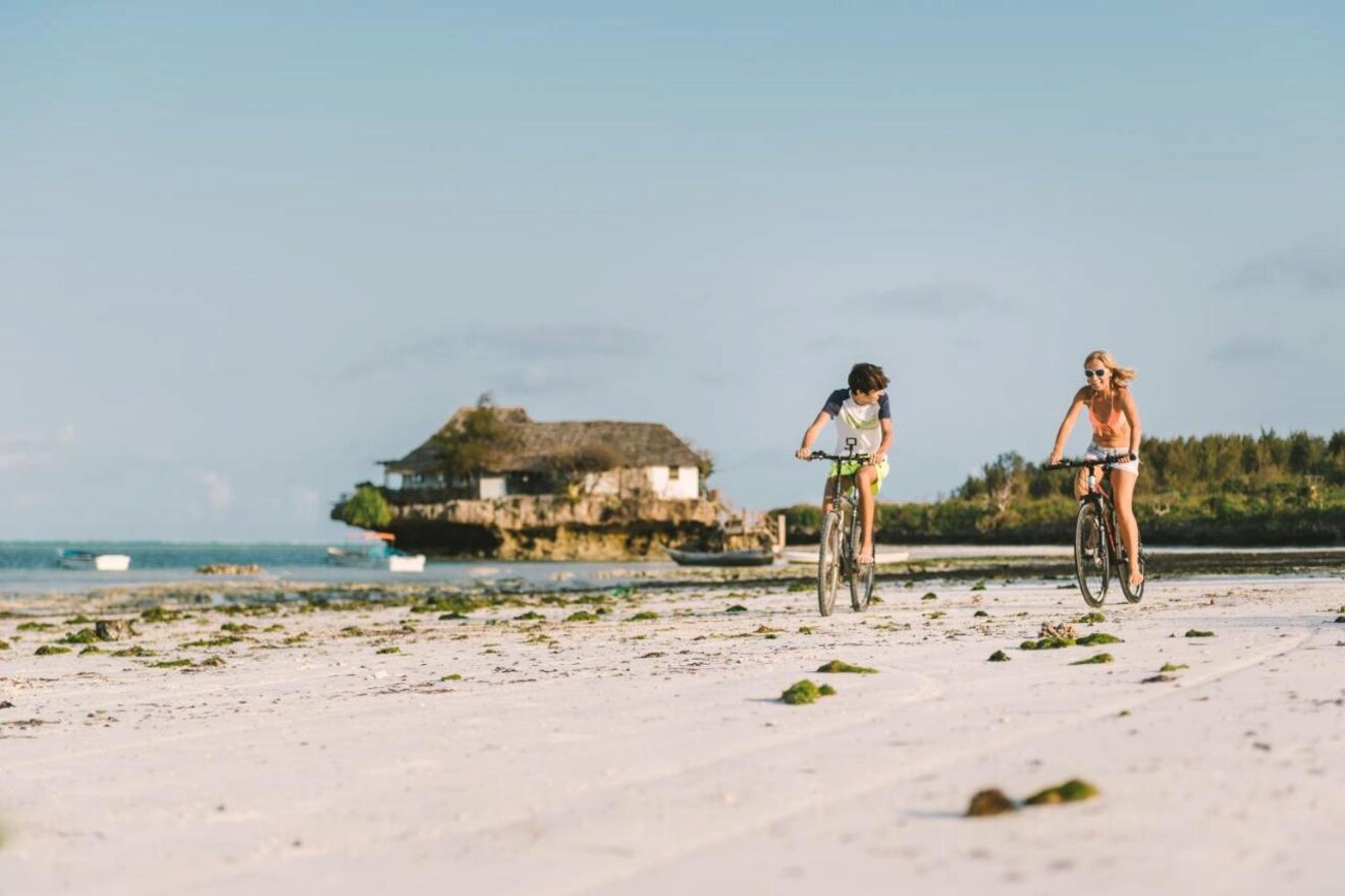 a girl and a boy cycling on the Pingwe beach in Zanzibar with the Rock restaurant in the background