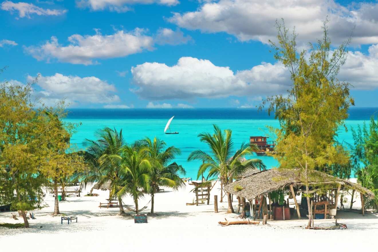 A white sand beach with palm trees and straw huts and a sailboat in the water in the background in Zanzibar