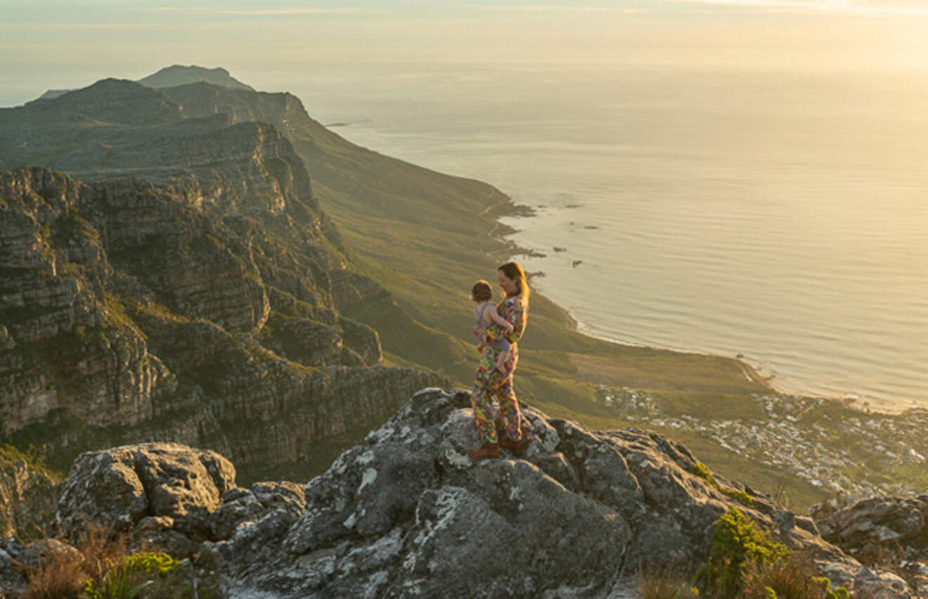 a woman holding a child standing on a rock with a landscape view of the mountains and the ocean