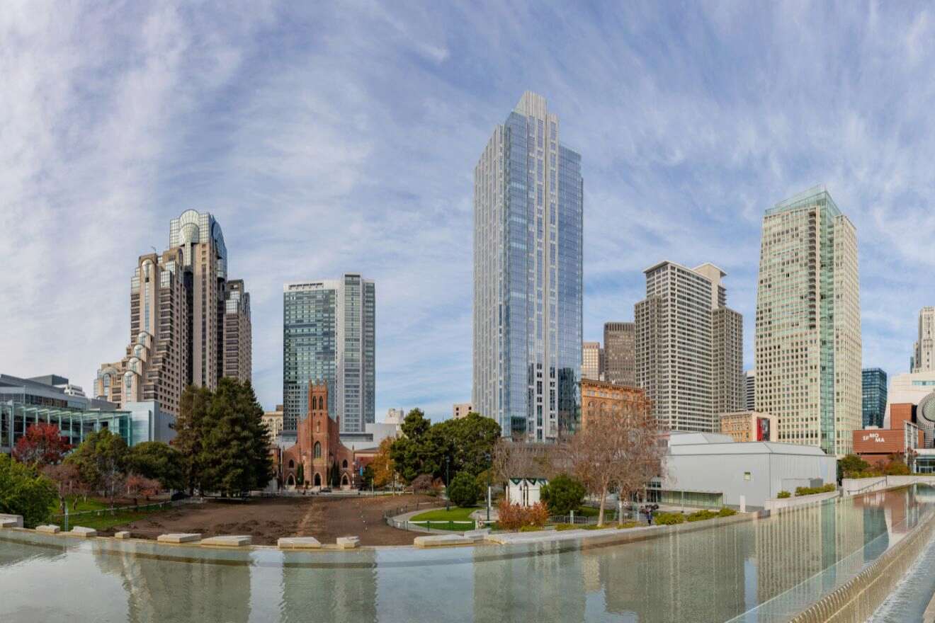 A panoramic view of the Soma and South Beach area in San Francisco, showcasing a mix of residential towers and the historical red-bricked church by a reflective waterfront park.