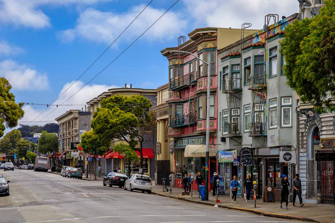 Colorful Victorian architecture lines a street in San Francisco with pedestrians and parked cars, set against a backdrop of lush trees and overcast skies