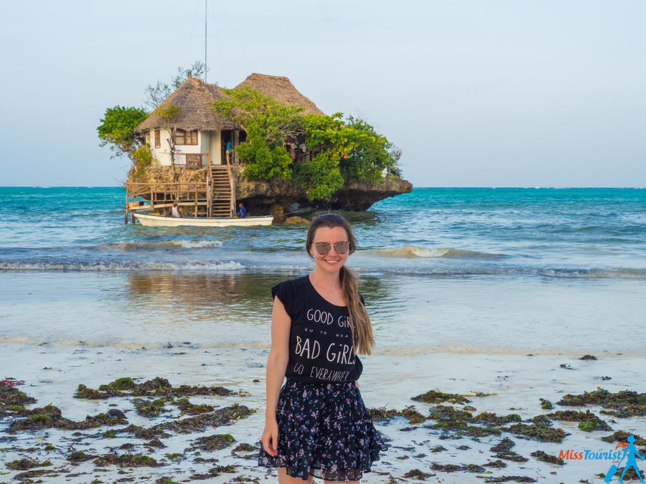 A girl in black outfit and sunglasses posing on the beach with the Rock restaurant in Zanzibar in the background