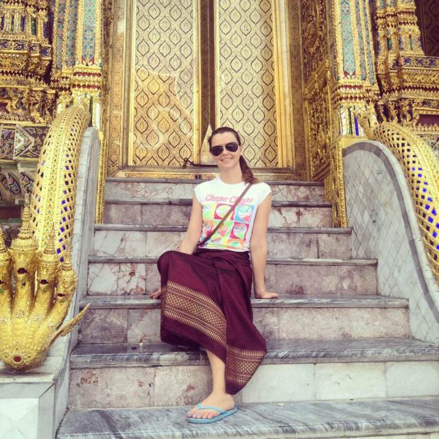 Yulia, the writer of the post poses on the steps of a golden-decorated temple in Bangkok, wearing a casual outfit with a traditional Thai sarong.