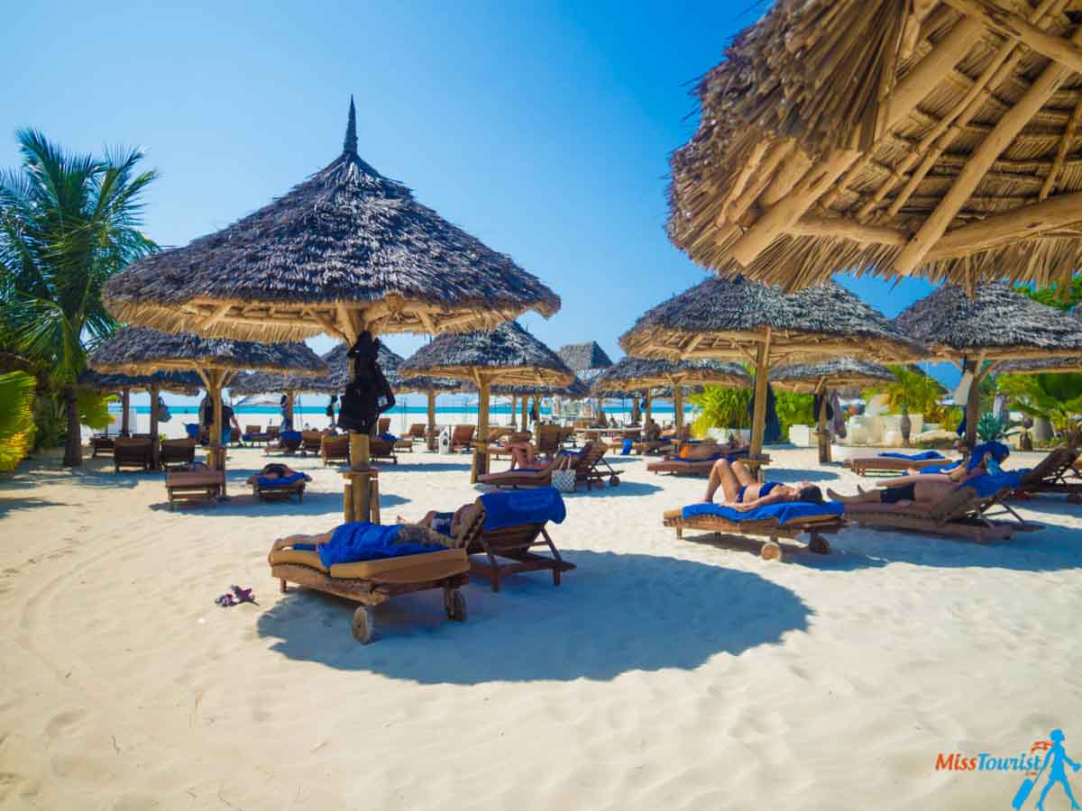 People laying on lounge chairs under straw umbrellas on a sandy beach in Zanzibar