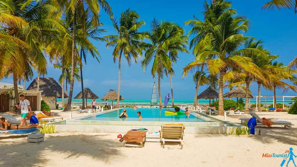 People swimming and relaxing on lounge chairs around a hotel pool surrounded by palm trees by the beach in Zanzibar