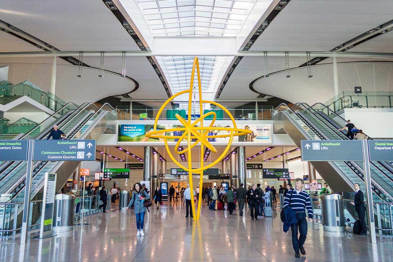 Interior of Dublin Airport featuring travelers, a striking yellow abstract sculpture, informational signage, escalators, and the airy architecture of the terminal with a glass ceiling