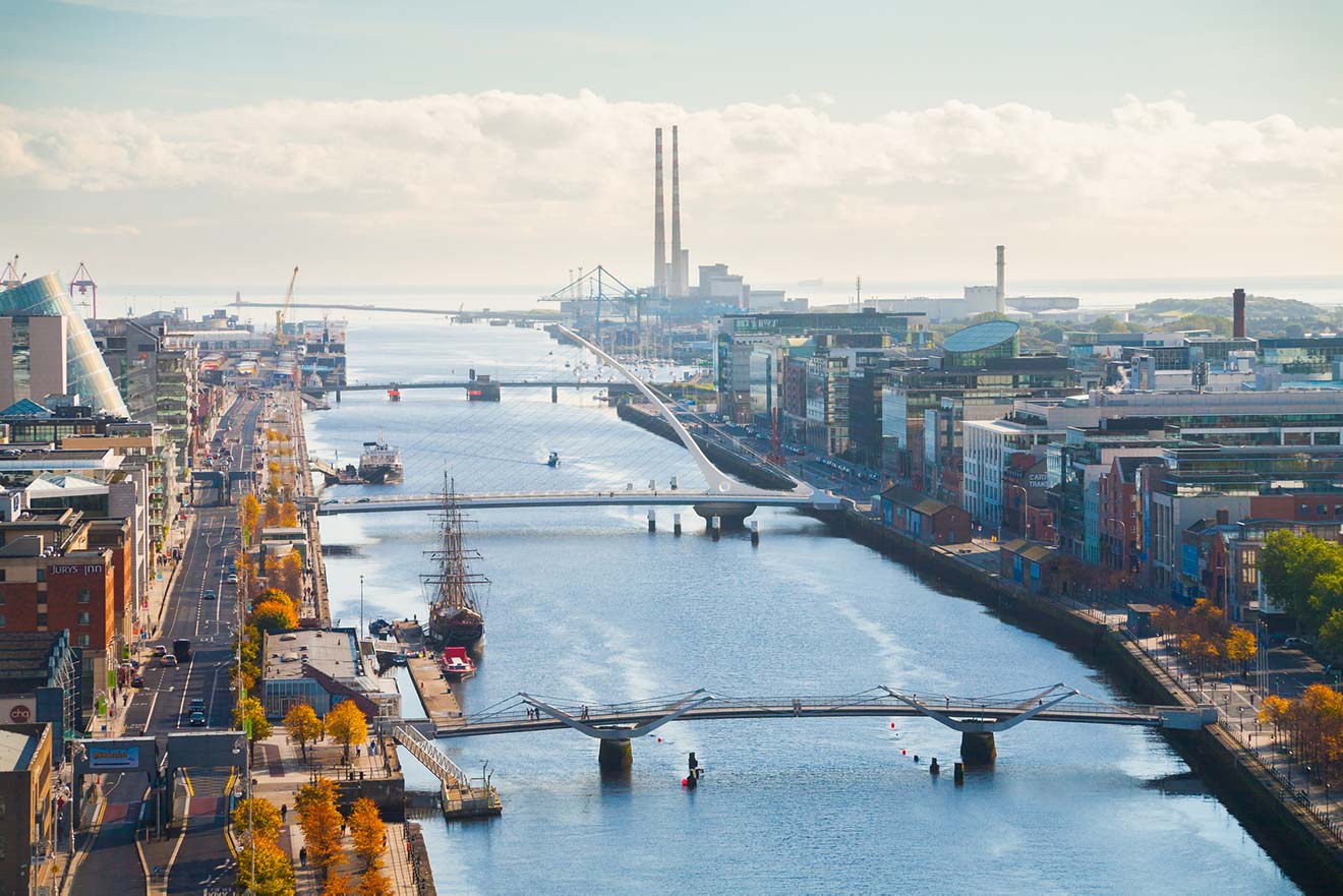 Aerial view of the River Liffey flowing through Dublin, with the Samuel Beckett Bridge in the foreground, leading the eye towards the docklands and industrial chimneys in the distance, on a clear day.