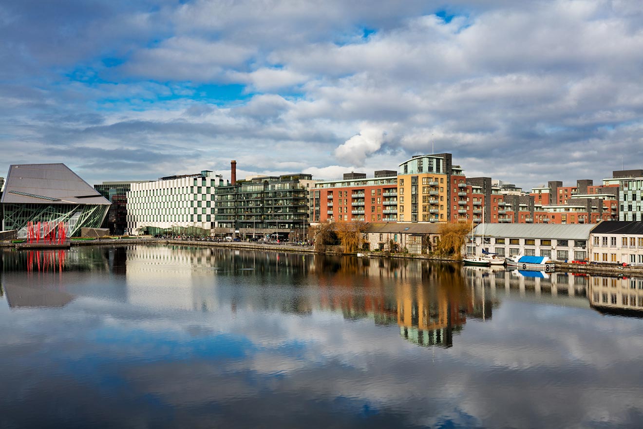 Modern urban waterfront with a mix of architectural styles reflected in the calm waters, featuring the geometric facade of the Bord Gáis Energy Theatre in Dublin under a dynamic sky with fluffy clouds