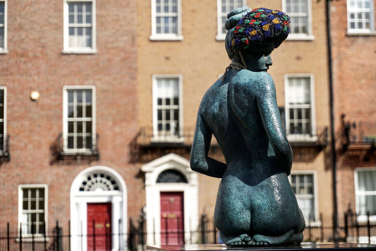 A bronze statue of a seated woman with a beaded hat, set against a backdrop of Georgian-style red brick buildings with white-trimmed windows and classic red doors in Dublin.