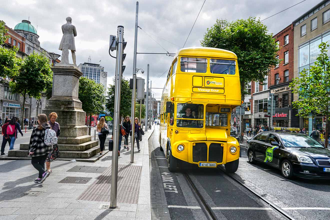 A vibrant yellow vintage double-decker bus, marked 'City Tours Vintage Tour', drives through a bustling urban street with pedestrians, modern vehicles, and a statue on a pedestal under a partly cloudy sky in Dublin