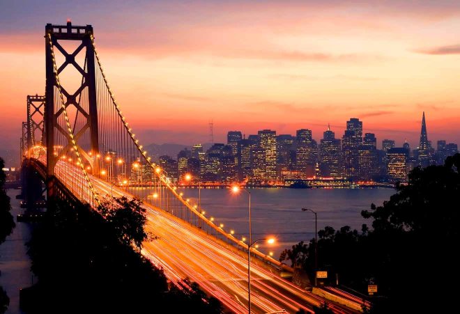 The Bay Bridge leading into the illuminated San Francisco skyline at dusk, with long exposure traffic lights tracing a vibrant path into the city under a twilight sky