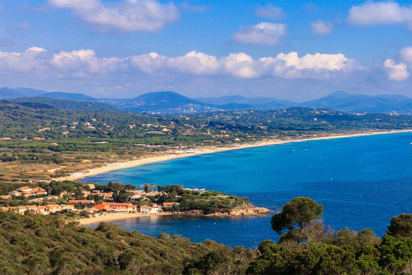 A scenic view of a bay in Saint-Tropez with a clear blue sea, sandy beach, green hills in the background, and a blue sky above