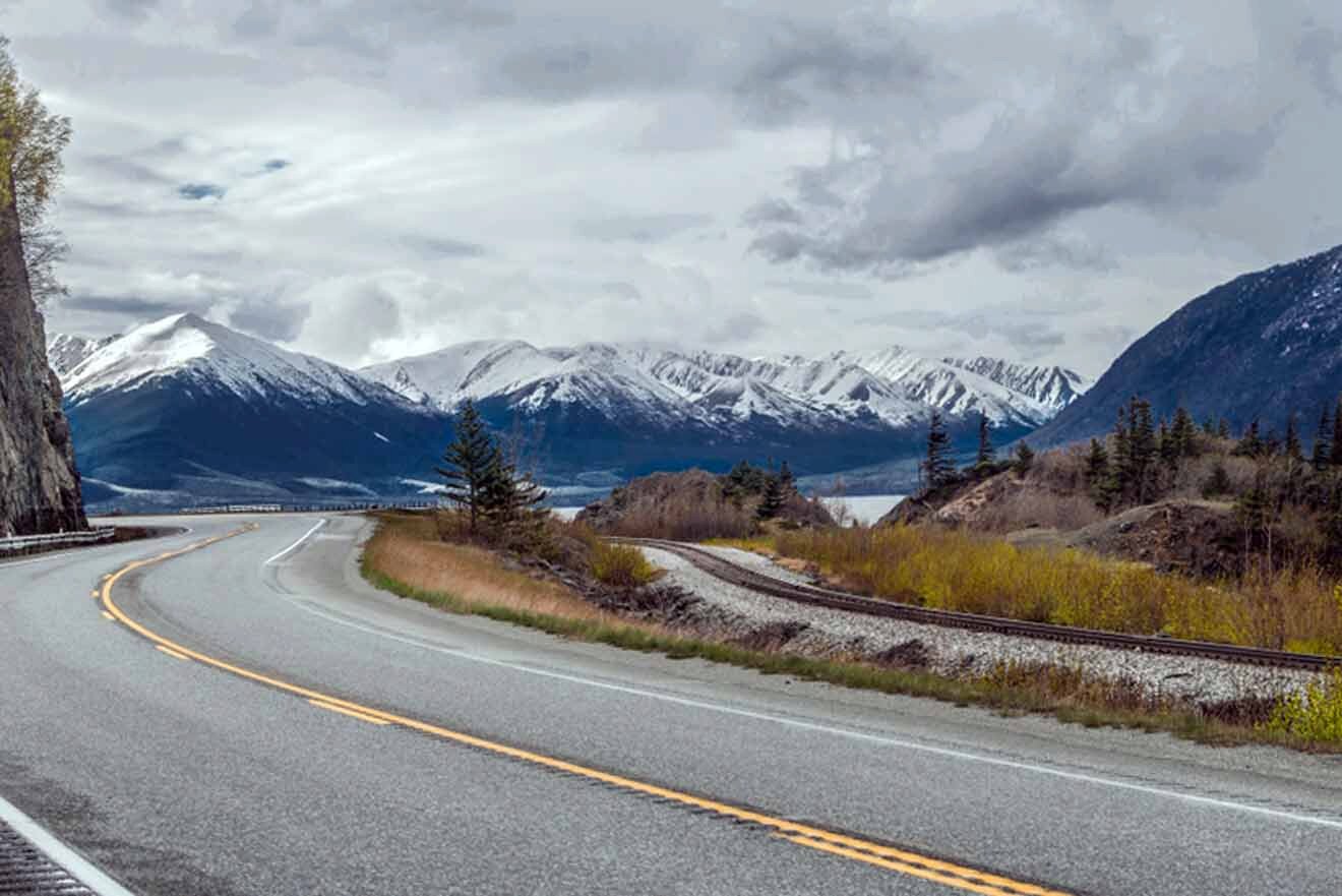 A scenic view of an empty road curving beside a railway track, with snow-capped mountains in the distance and a partly cloudy sky.