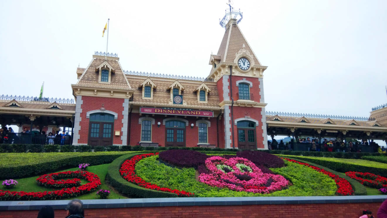 The entrance to Hong Kong Disneyland with a floral Mickey Mouse design in the foreground and the iconic Disneyland railway station in the background.