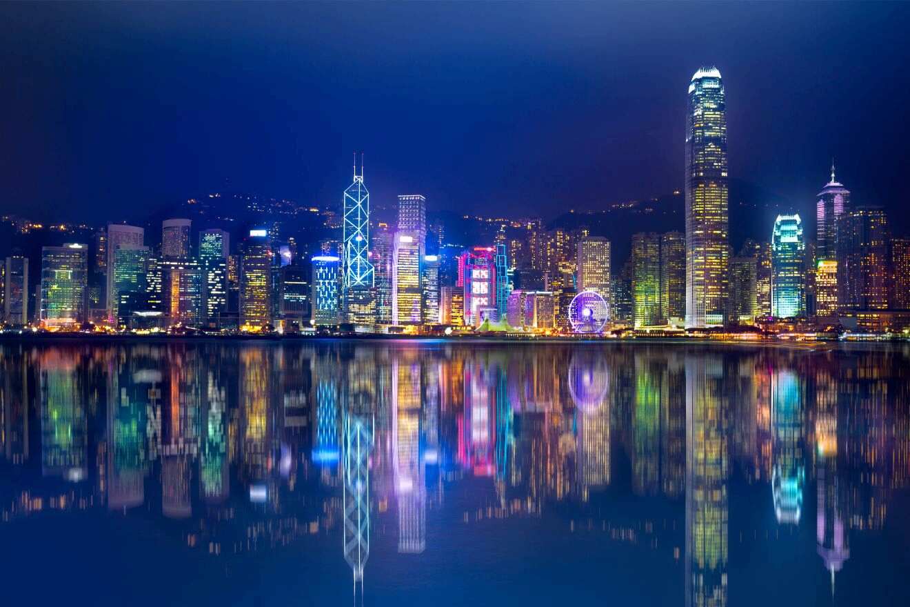 A panoramic night view of Hong Kong's illuminated skyline reflected in the calm waters of Victoria Harbour, showcasing a vibrant cityscape.