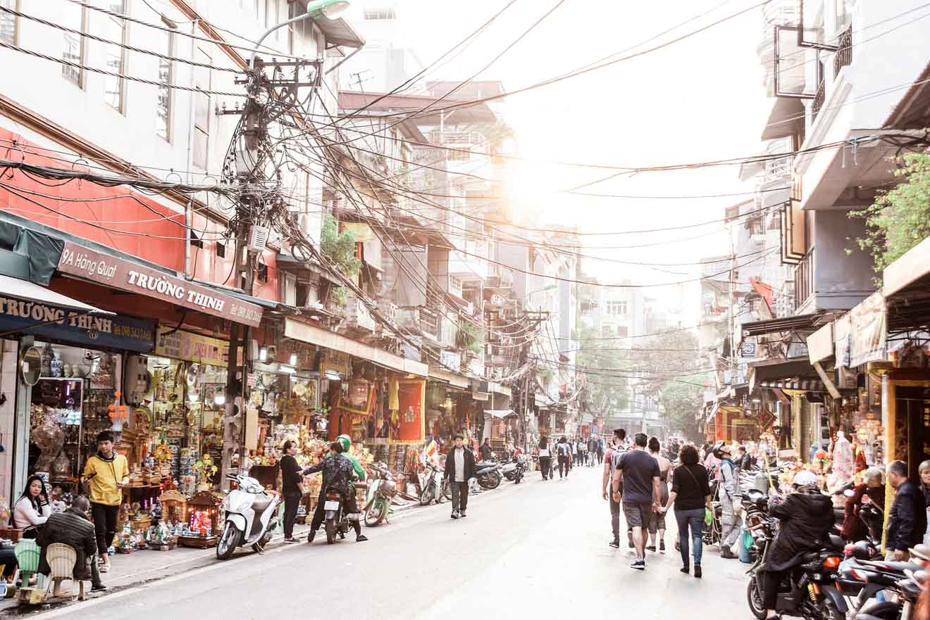 Busy daytime street scene in Hanoi with pedestrians and motorbikes among traditional shops and tangled overhead wires, highlighting the city's energetic atmosphere