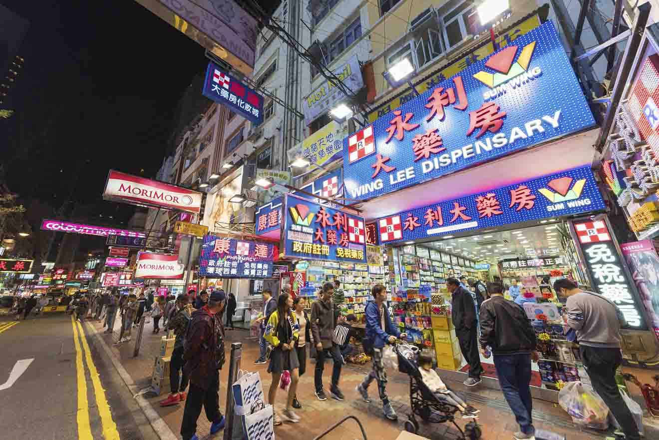 A vibrant street scene in Hong Kong with bright neon signs in various languages, pedestrians, and a plethora of small shops and pharmacies.