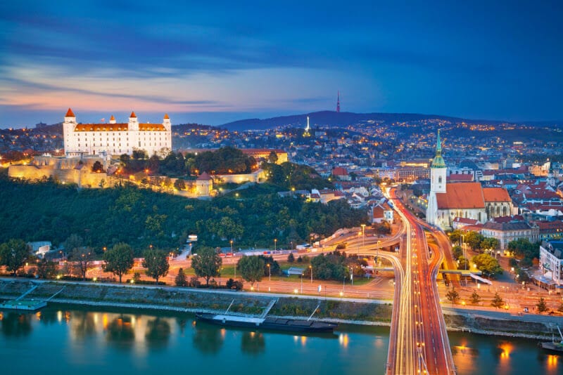 view of castle on hill and church beside river with bridge going over it all lit up at night