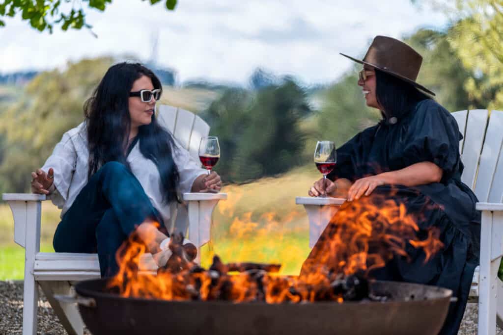 two women sitting around firepit drinking wine