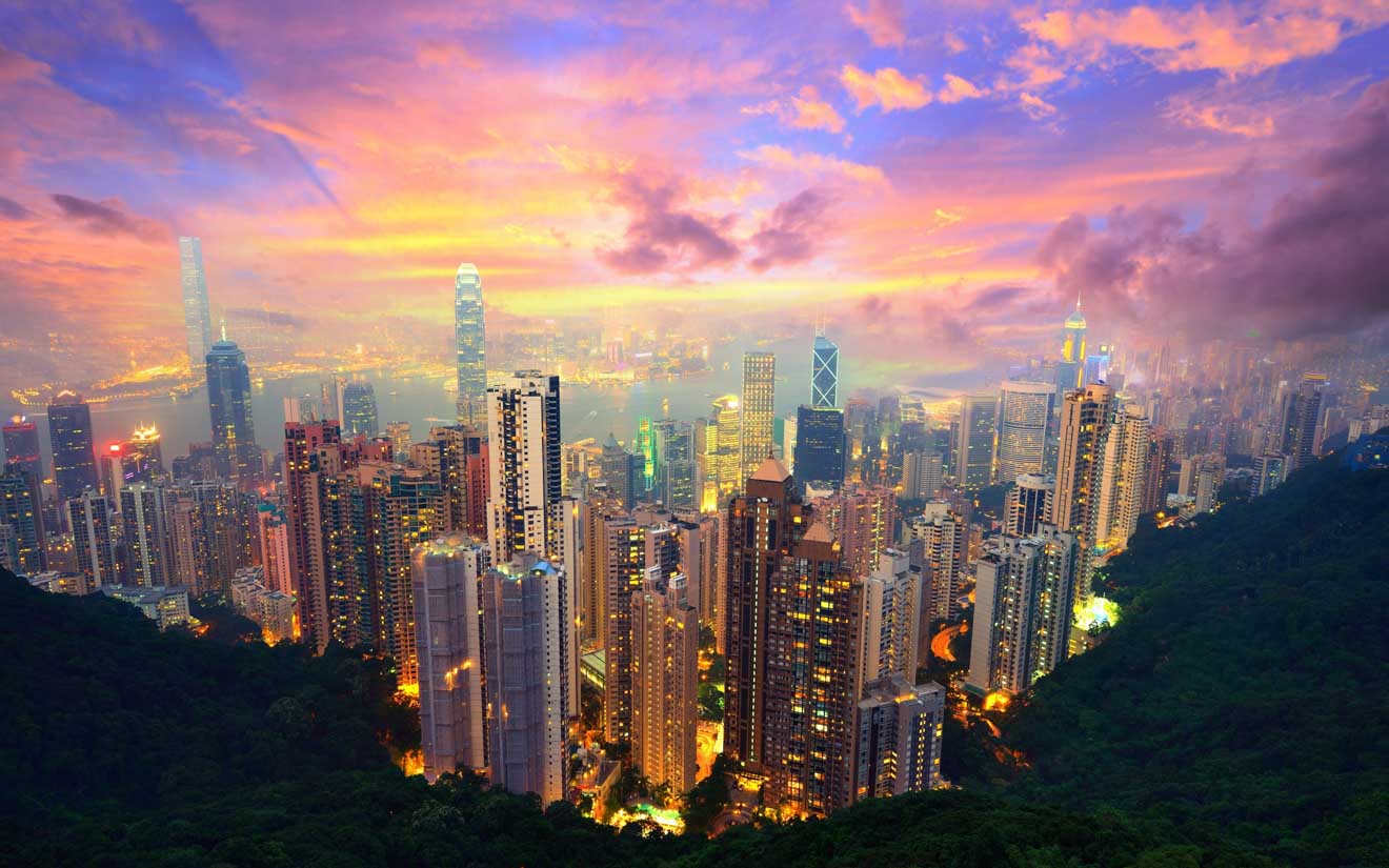 A panoramic view of Hong Kong's skyline with towering skyscrapers illuminated at dusk against a vibrant sunset sky, showcasing the city's dense urban architecture.