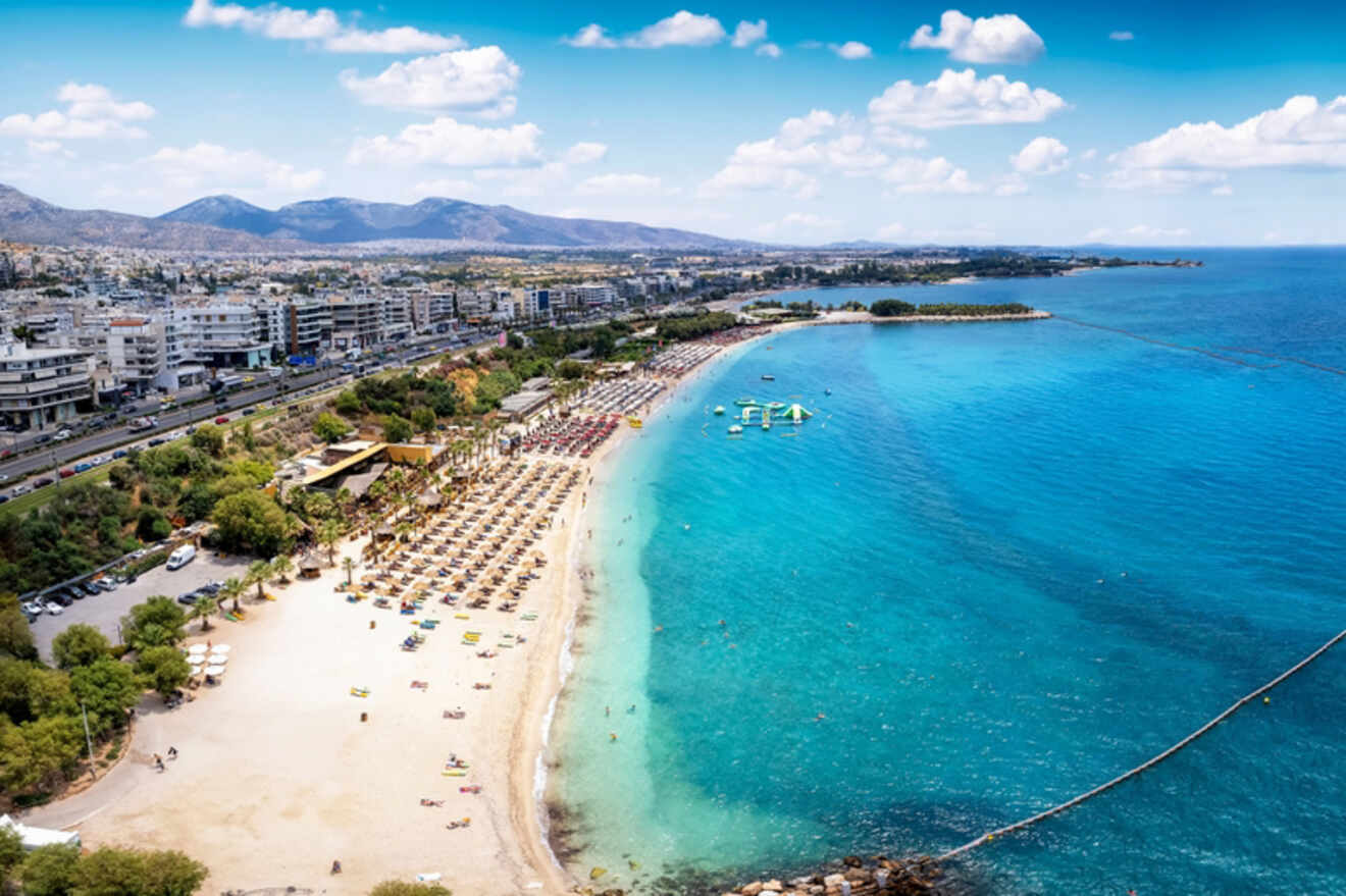 A curving beachfront in Athens with rows of umbrellas and sunbathers, a floating play area in the sea, and the cityscape in the background