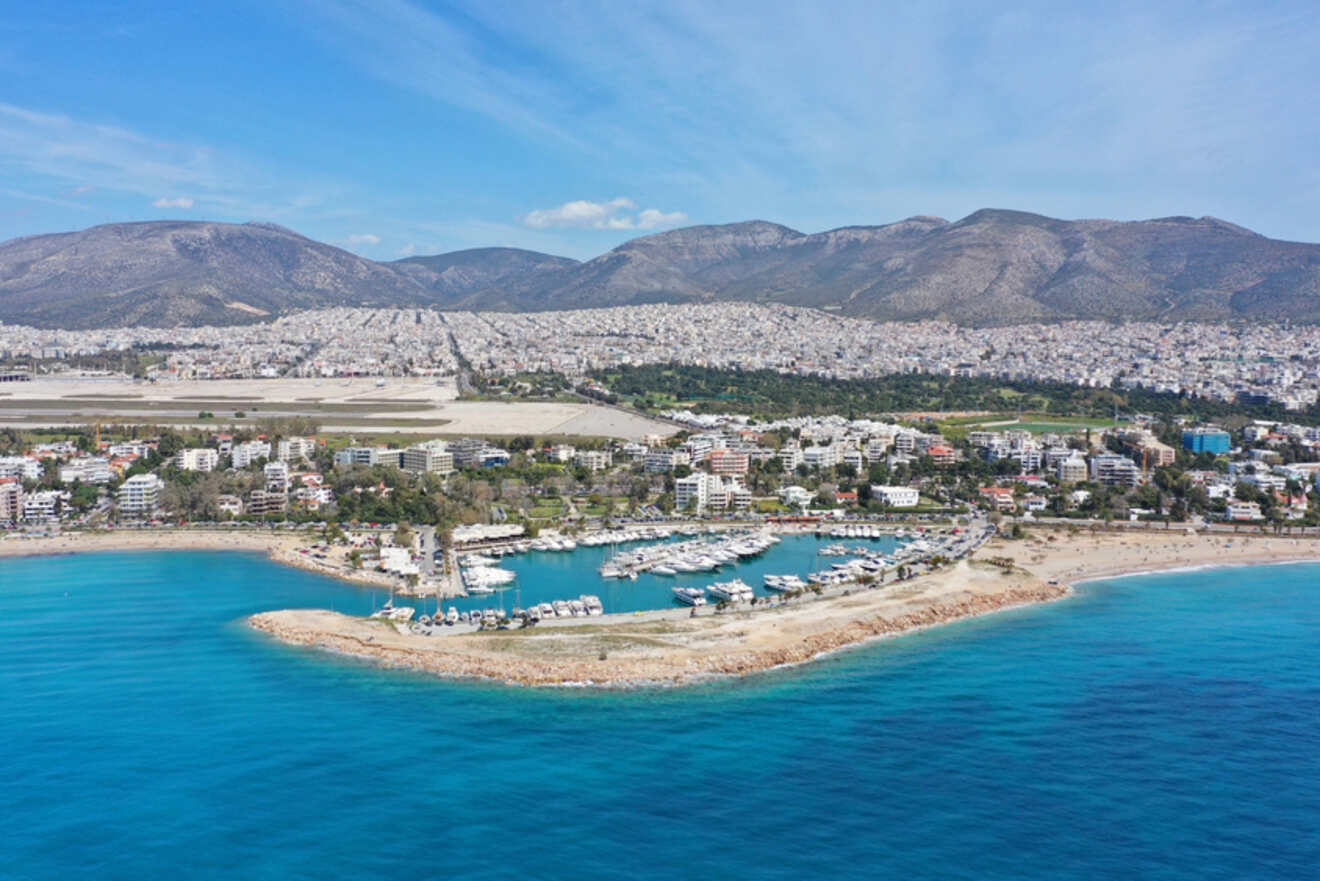 Aerial view of a coastal Athens neighborhood with boats docked in a marina, clear blue waters, and a backdrop of densely packed buildings and hills