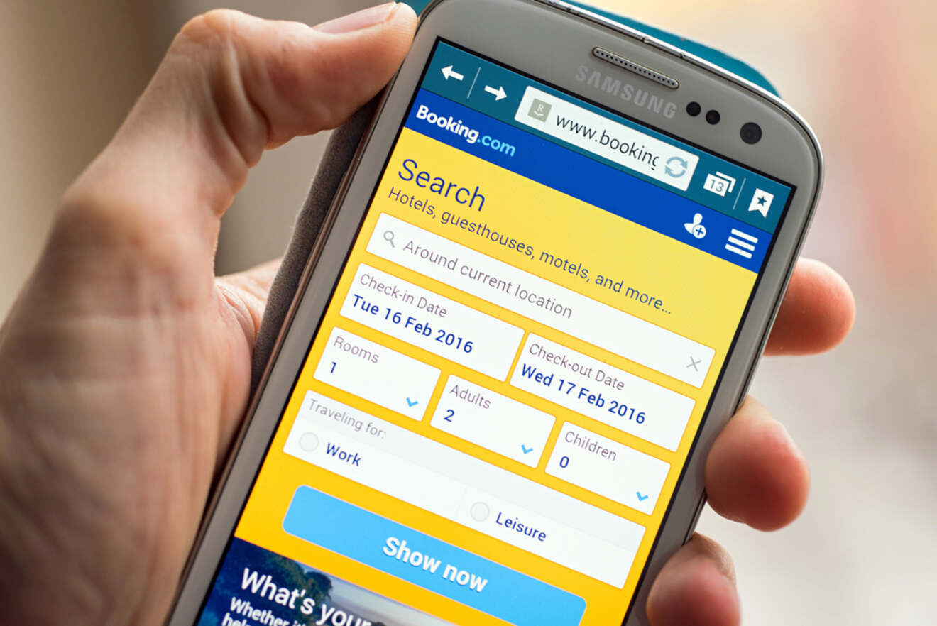 Hand holding a Samsung smartphone with the Booking.com mobile site displayed, showing the search interface for hotels, guesthouses, and motels. The screen indicates options for check-in and check-out dates, number of rooms and adults