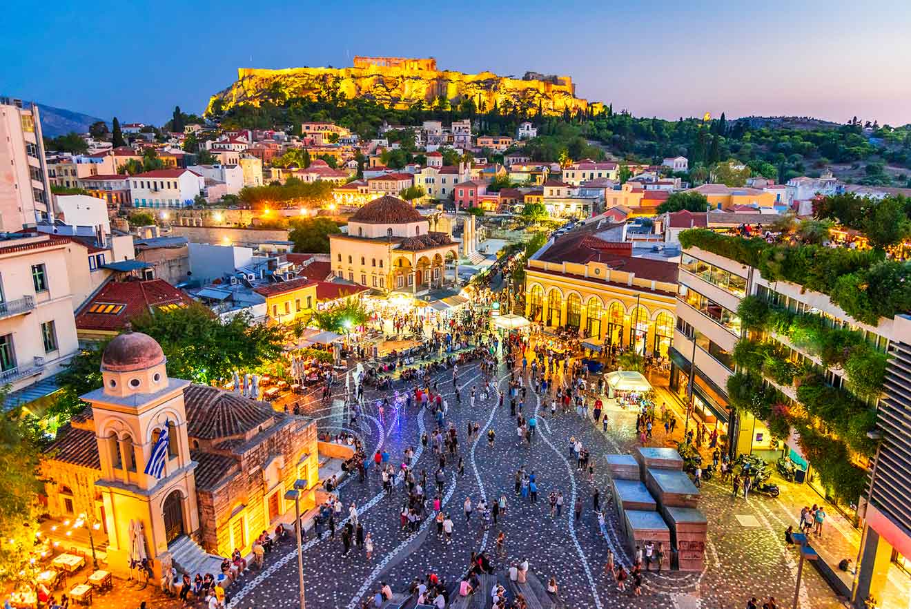 Twilight view over a bustling Athens square filled with people, market stalls, and lined with historic architecture, with the illuminated Acropolis in the distance