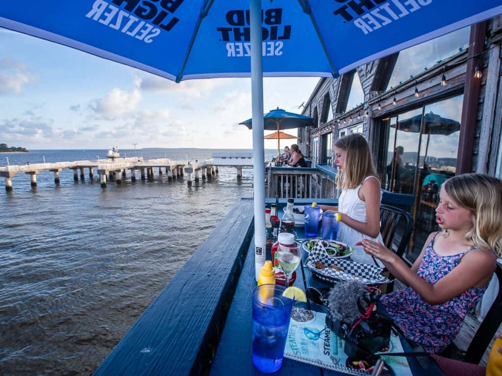 Two girls eating dinner on the deck of a restaurant overlooking the water.